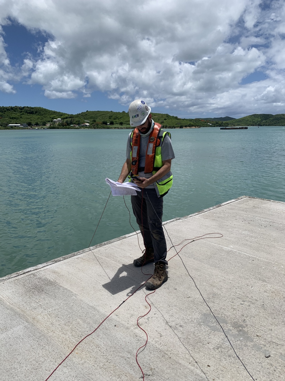 A VCS employee in a safety vest and helmet stands on a waterfront dock, holding papers and a multimeter with cables extending into the water.