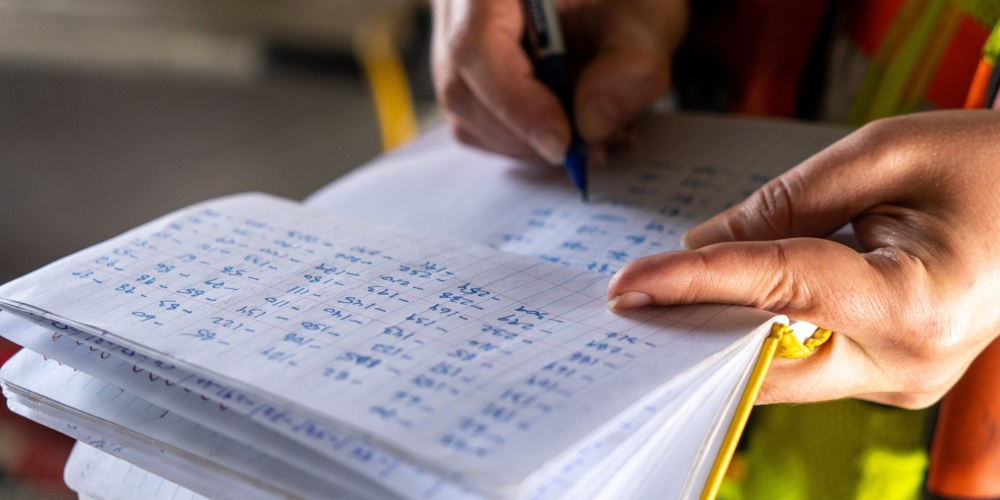 person writing in a notebook with structural data written on it in columns.
