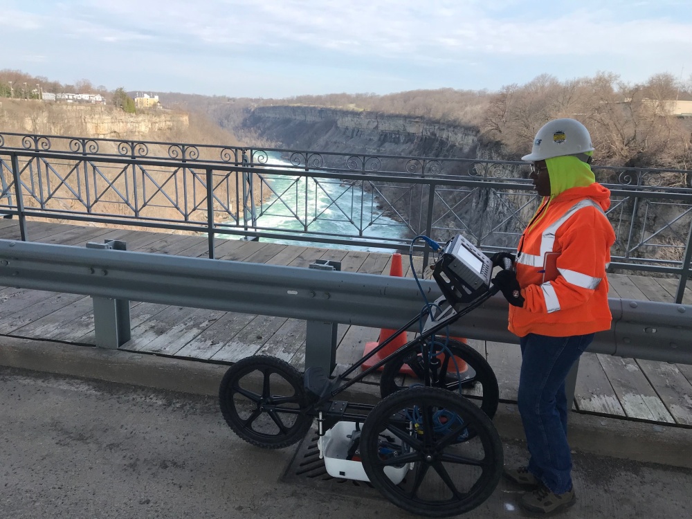 Person standing behind a GPR device on a tri-wheeled cart over looking a river from a bridge deck .