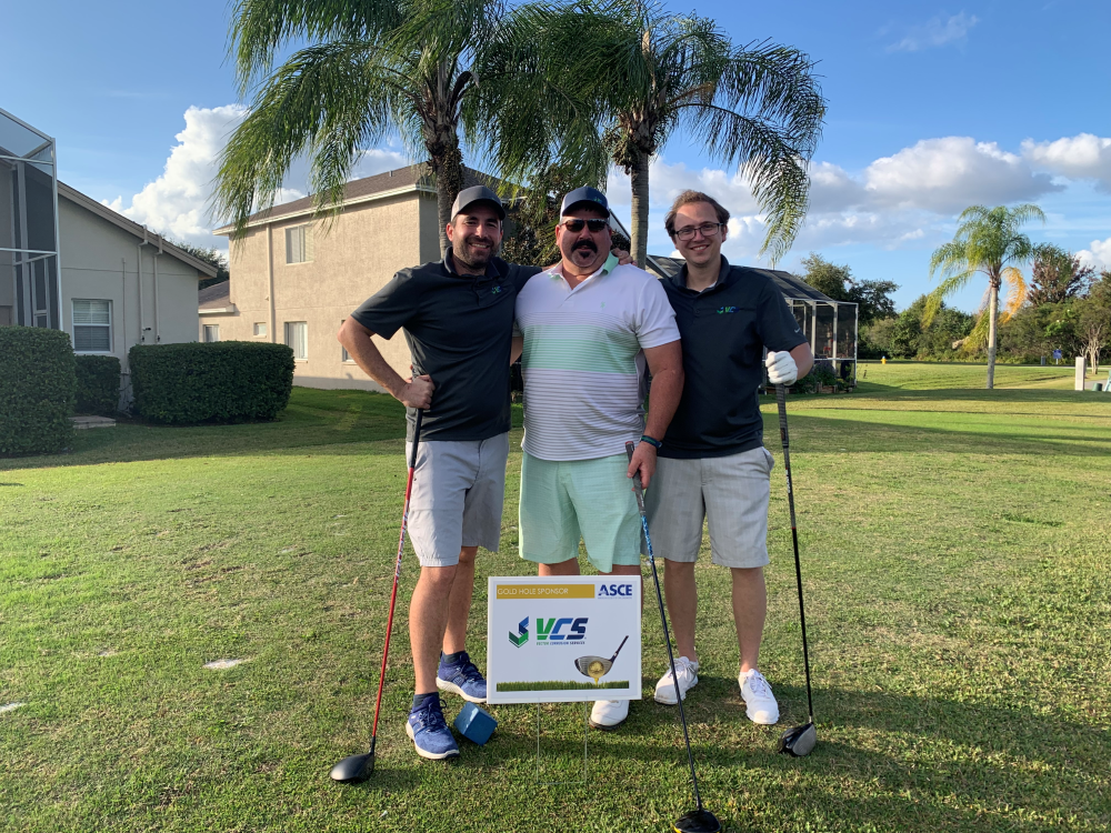 Group of three men standing behind a VCS Engineering sign at a golf tournament, smiling and holding golf clubs.